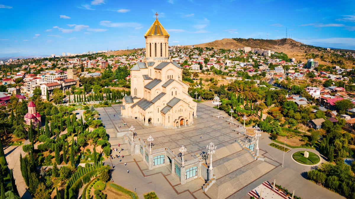 Aerial view of the Holy Trinity Cathedral (Sameba) in Tbilisi, Georgia, featuring its iconic golden dome, landscaped grounds, and panoramic city skyline under a clear blue sky.