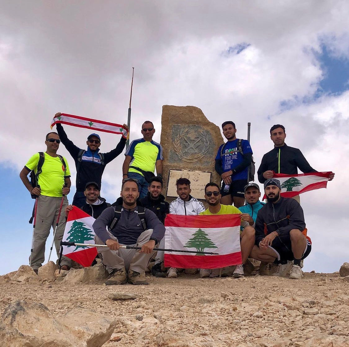 Hiking group posing with Lebanese flags at the summit marker of Qurnat As Sawda, Lebanon's highest peak at 3,088 meters, during a mountain trekking adventure.
