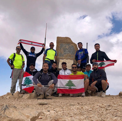 Hiking group posing with Lebanese flags at the summit marker of Qurnat As Sawda, Lebanon's highest peak at 3,088 meters, during a mountain trekking adventure.