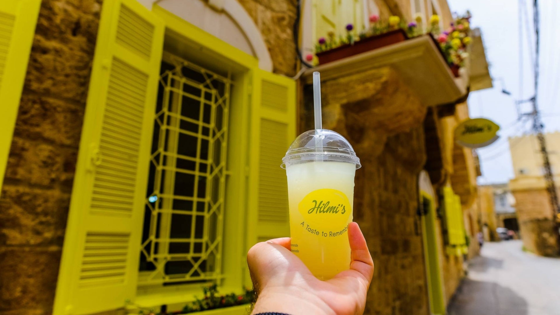 Hand holding a lemonade drink with 'Hilmi's' branding in front of a yellow building in Batroun Lebanon